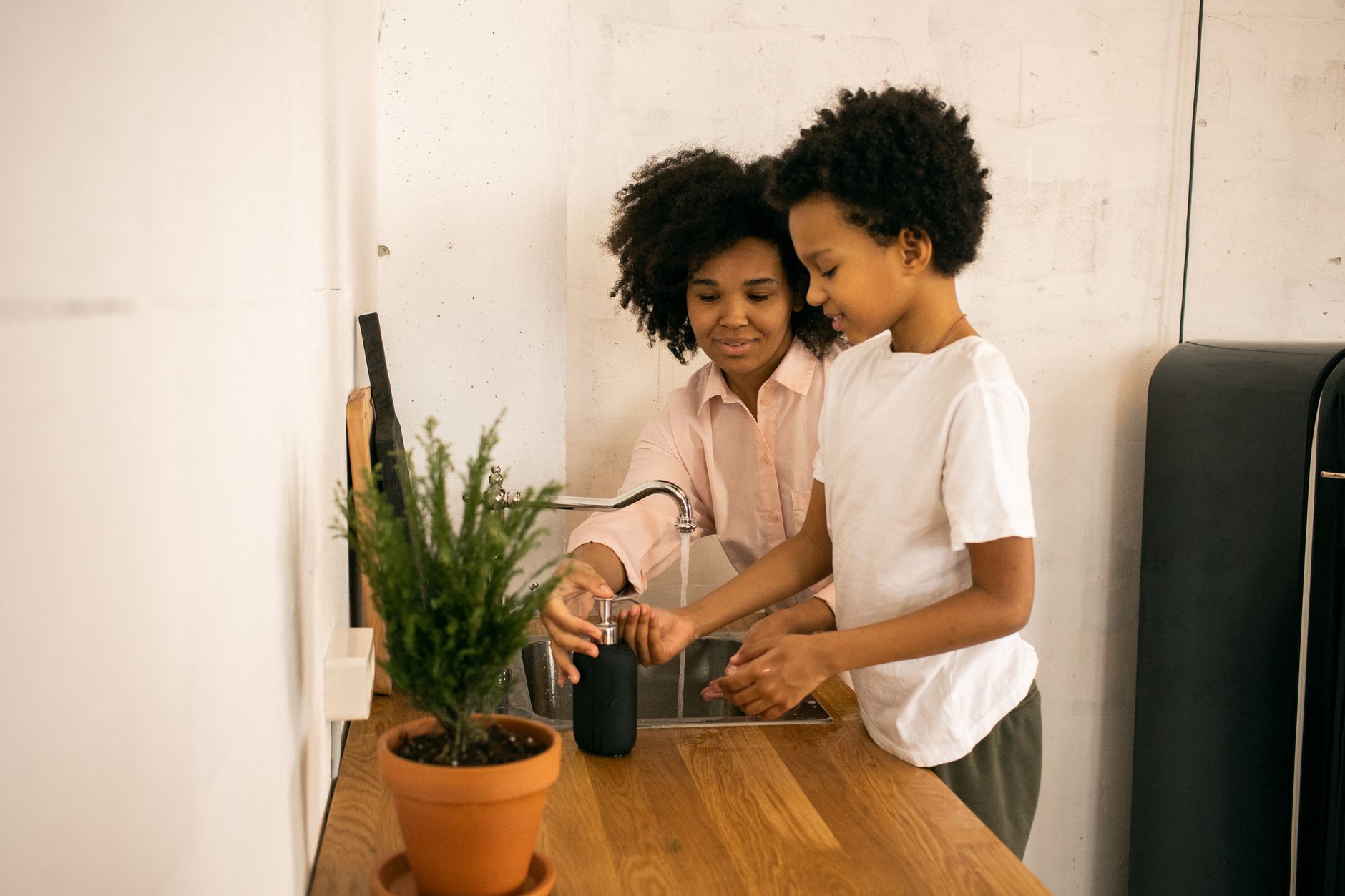 Glad African American mother with son applying soap on hands while washing hands near counter with green potted plant at home