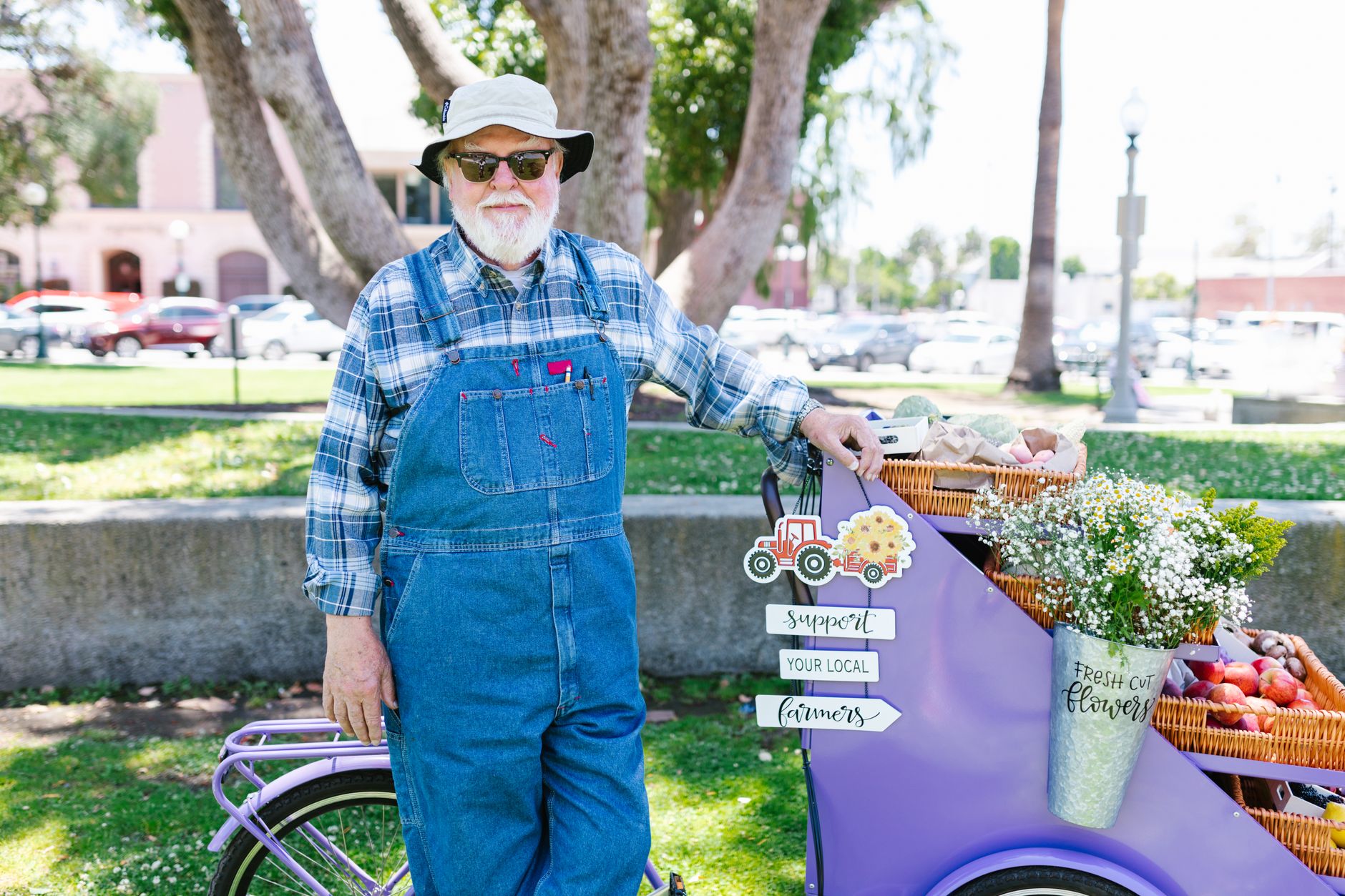 An elderly man selling fresh produce and flowers at an outdoor market. Support local farmers signage.