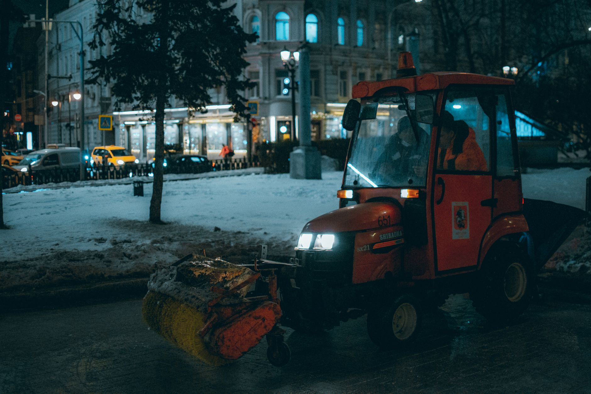 A red tractor clearing snow in an urban setting at night with lights and snow.