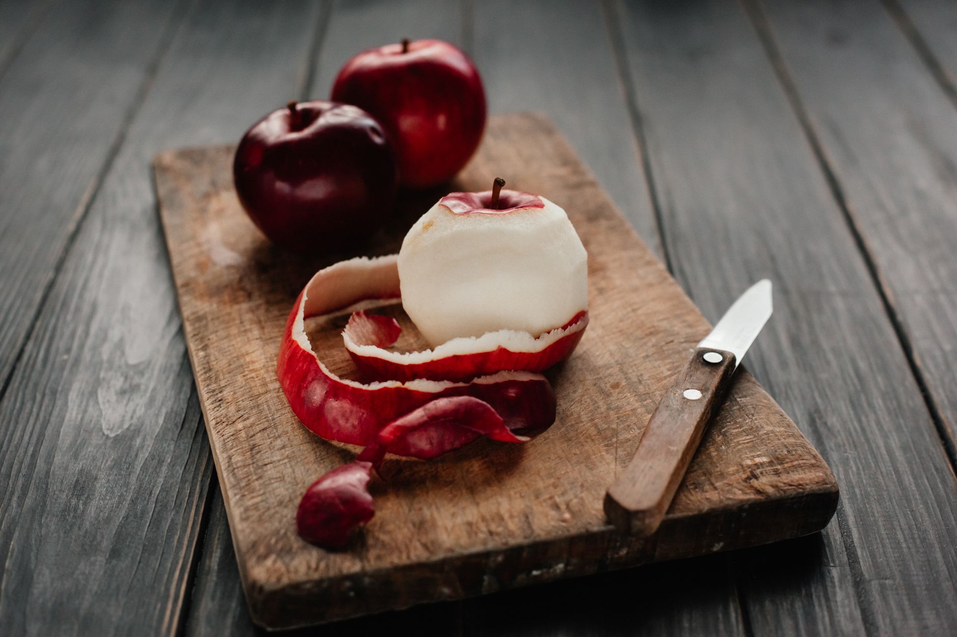 A peeled red apple with a knife on a rustic wooden chopping board.