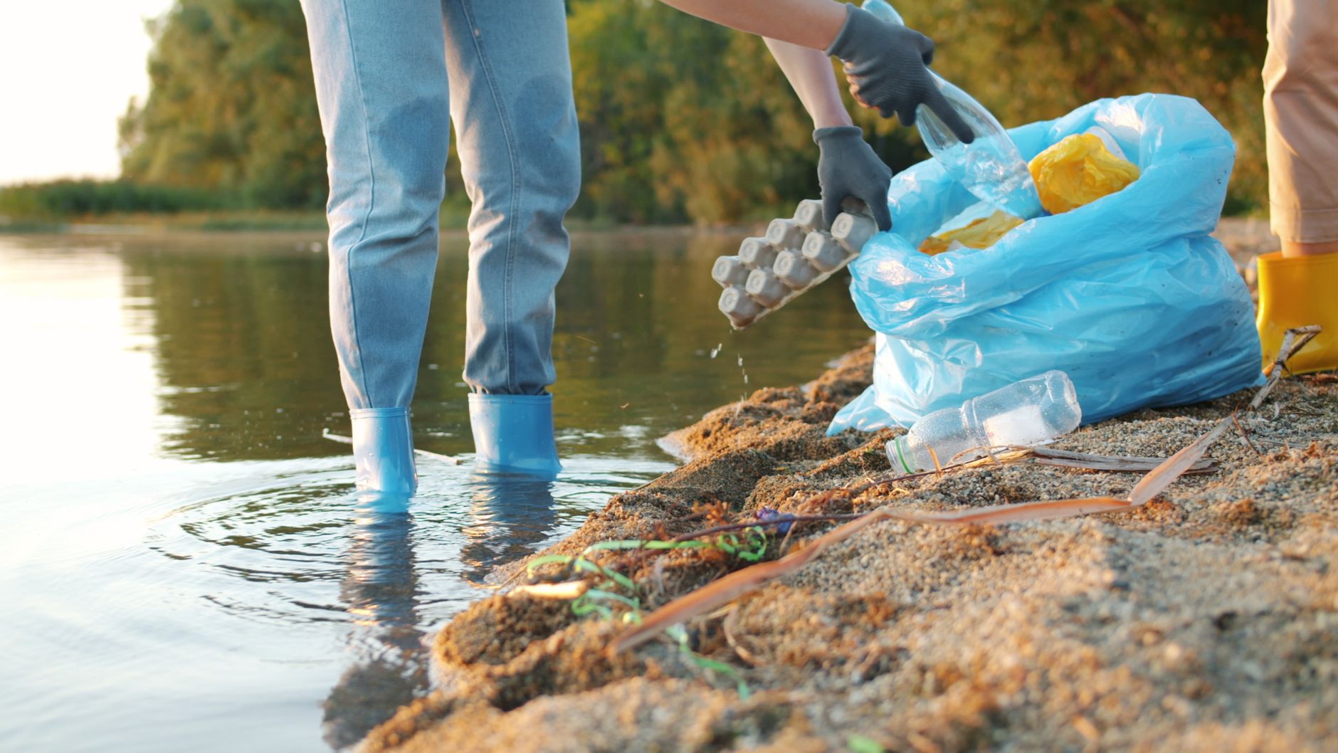 People cleaning a riverbank, collecting plastic waste in a blue bag with gloves.