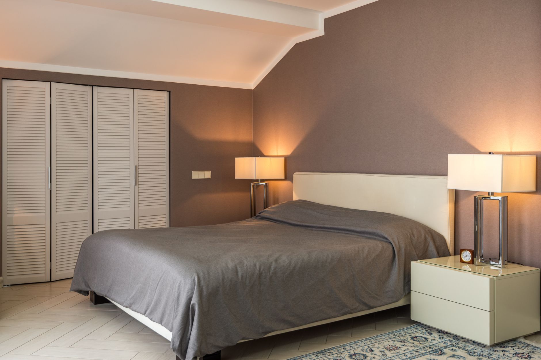 Comfortable bed covered with gray coverlet placed between bedsides with shining lamps near closet under geometric ceiling