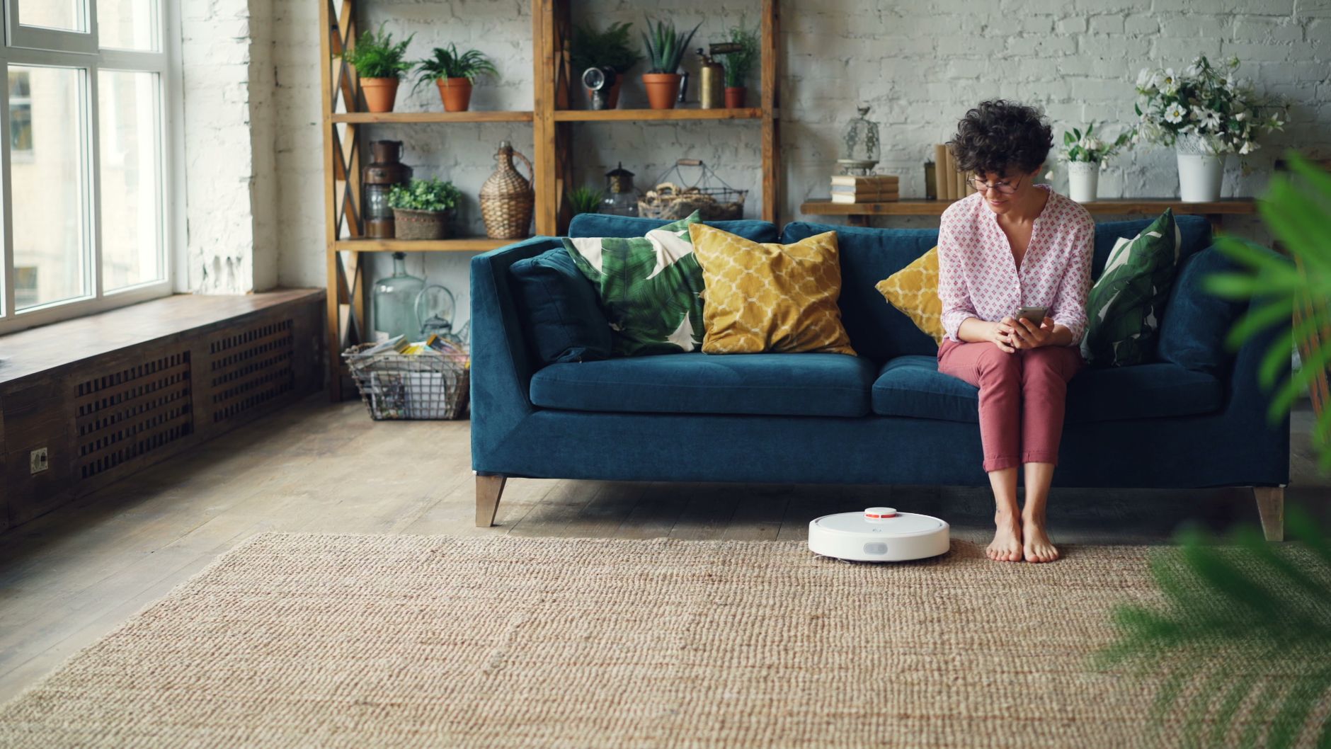 A woman sits on a sofa in a cozy living room with a robot vacuum on the floor.