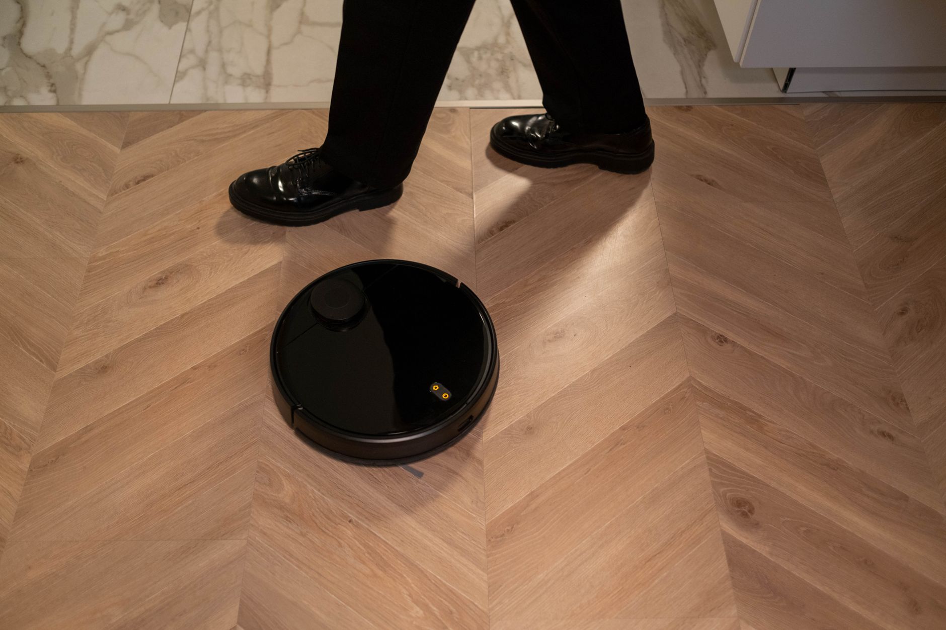 A modern robot vacuum cleaner navigating wooden floors while a person in black shoes walks past.