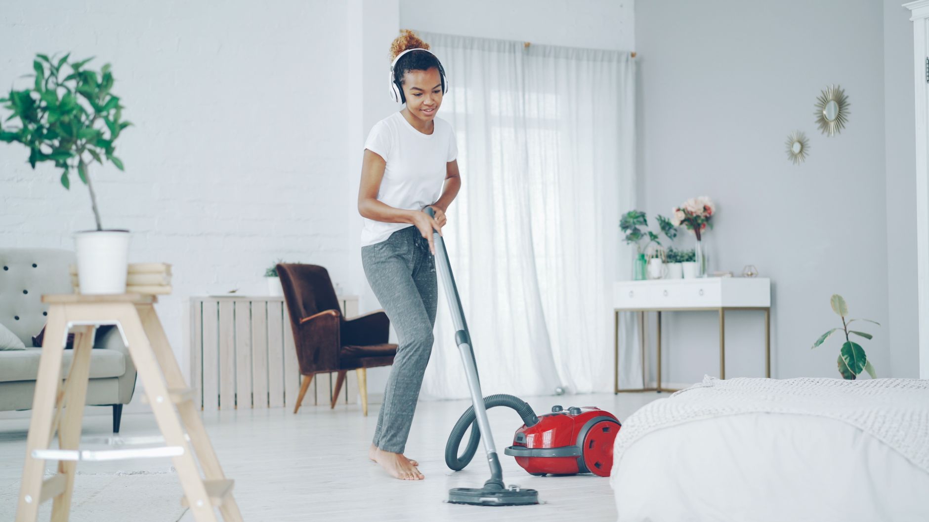 A woman in a cozy room uses a vacuum cleaner while enjoying music on headphones.