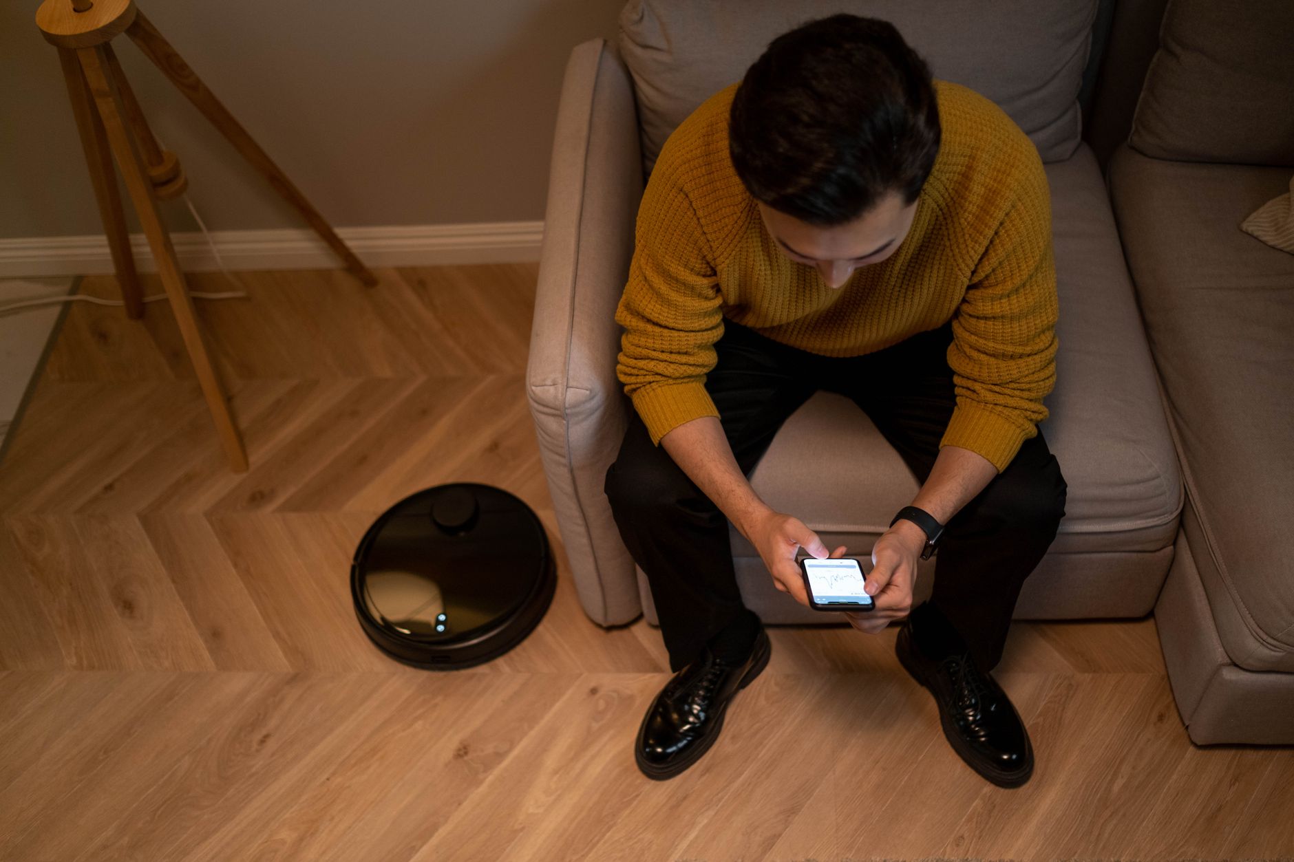 Man sits on sofa using a smartphone, next to a robotic vacuum cleaner in a modern living room.