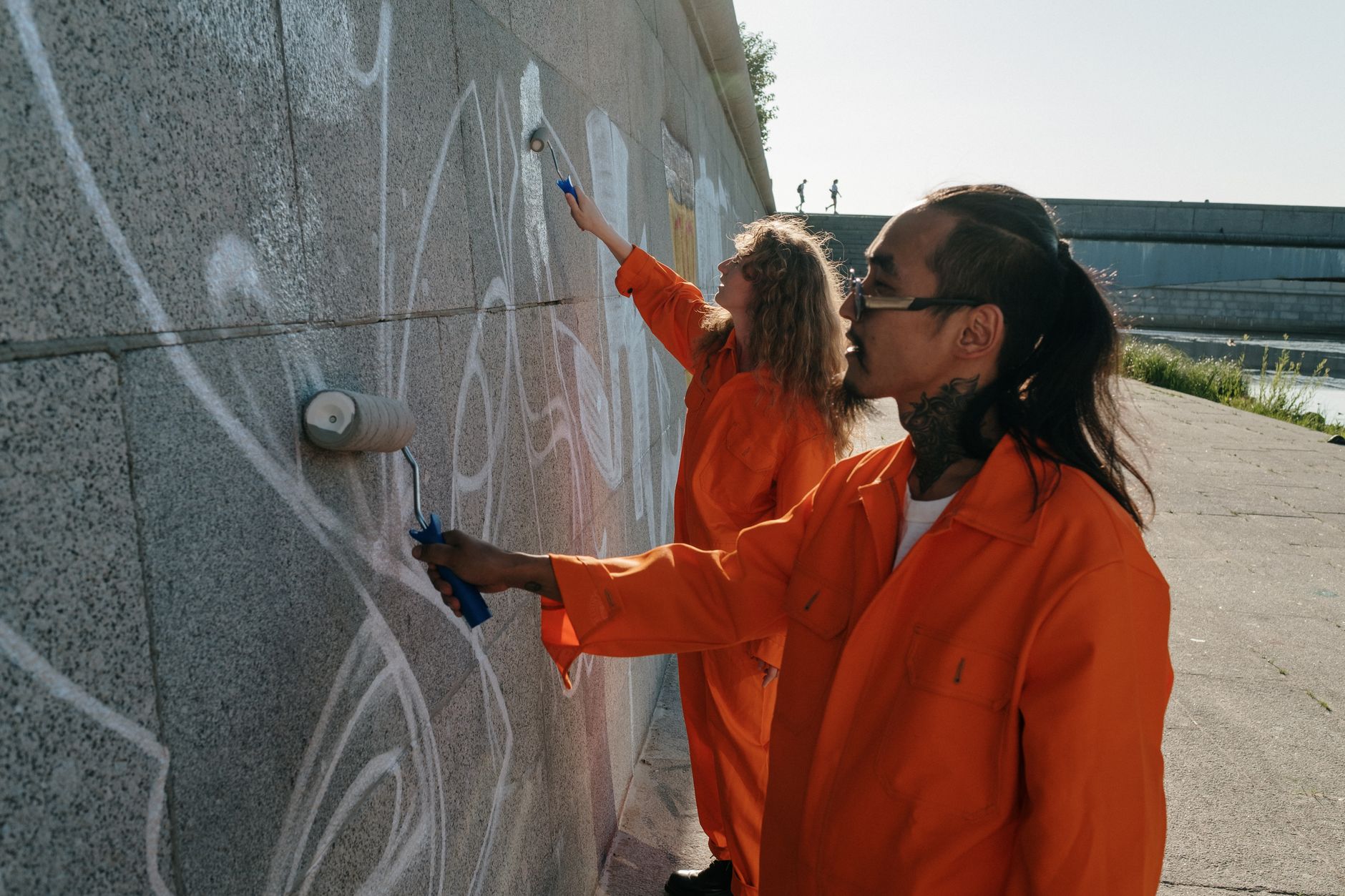 People in orange uniforms painting over graffiti on a concrete wall outdoors.