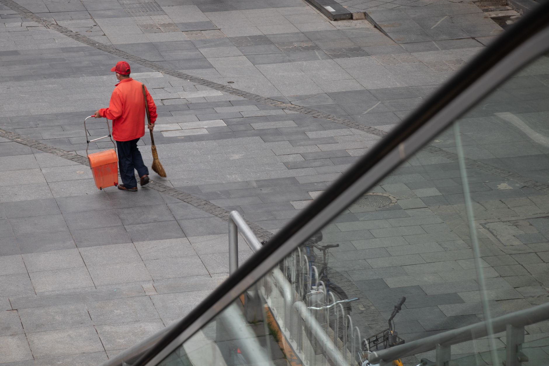 A street cleaner in a red jacket sweeping the urban pavement with a broom and cart.